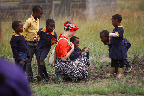 woman in red top and headdress crouches down as a child shows her something. They are surrounded by five other children 