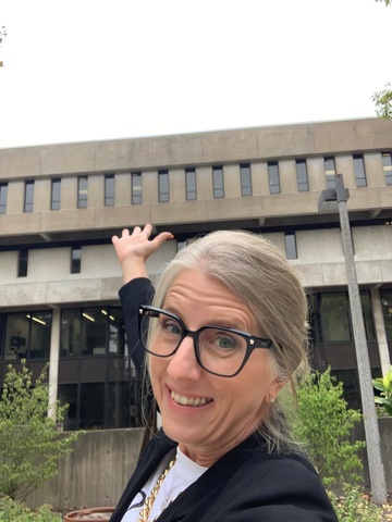 Selfie of woman in front of Nursing Building with her arm in the air pointed towards the building. 