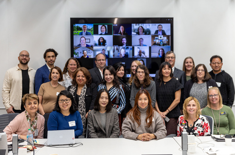 A group of about 20 researchers stand and sit for a group photo. They are in front of a tv that has remote participants shown in boxes. 