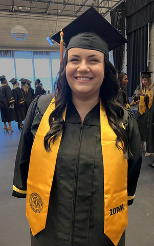 Woman in black graduation cap and gown with gold Iowa stole smiles at camera.